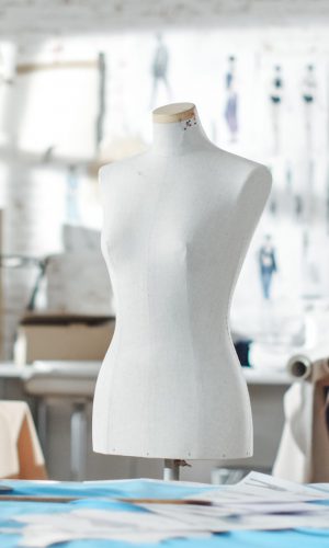 Shot of a Tailoring Mannequin that Stands in a Bright and Sunny Studio. Various Sewing Items and Colorful Fabrics Laying around, Mannequins Standing, and Sketches Pinned to the Wall.
